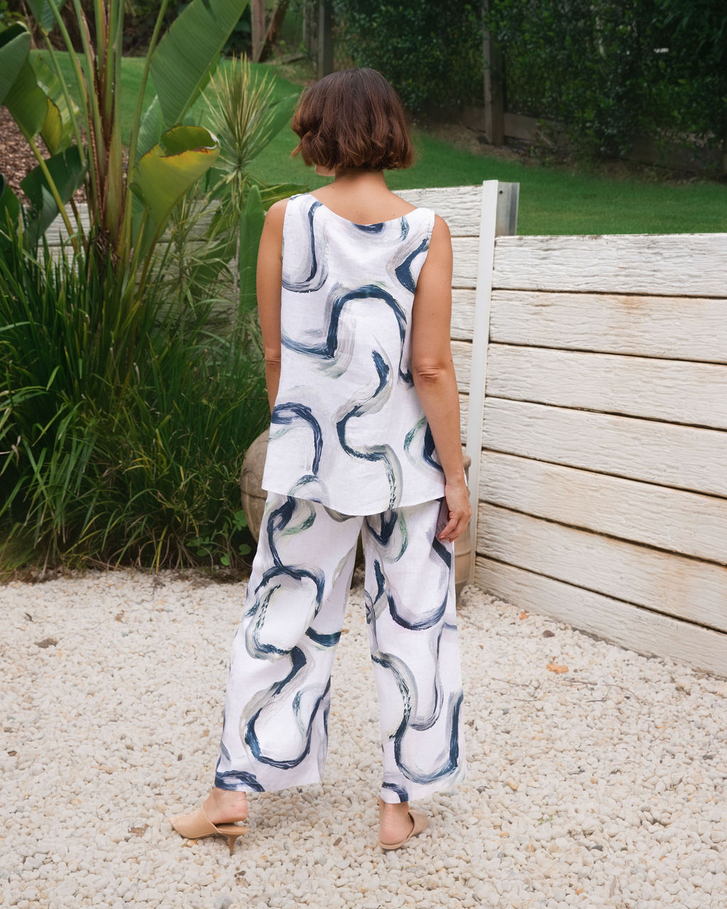 A woman wearing a Riverstone sleeveless white top and matching See Saw wide-leg pants with an abstract blue and gray brushstroke pattern, standing outdoors in a garden setting with large green plants, terracotta pots, and a light stone wall.