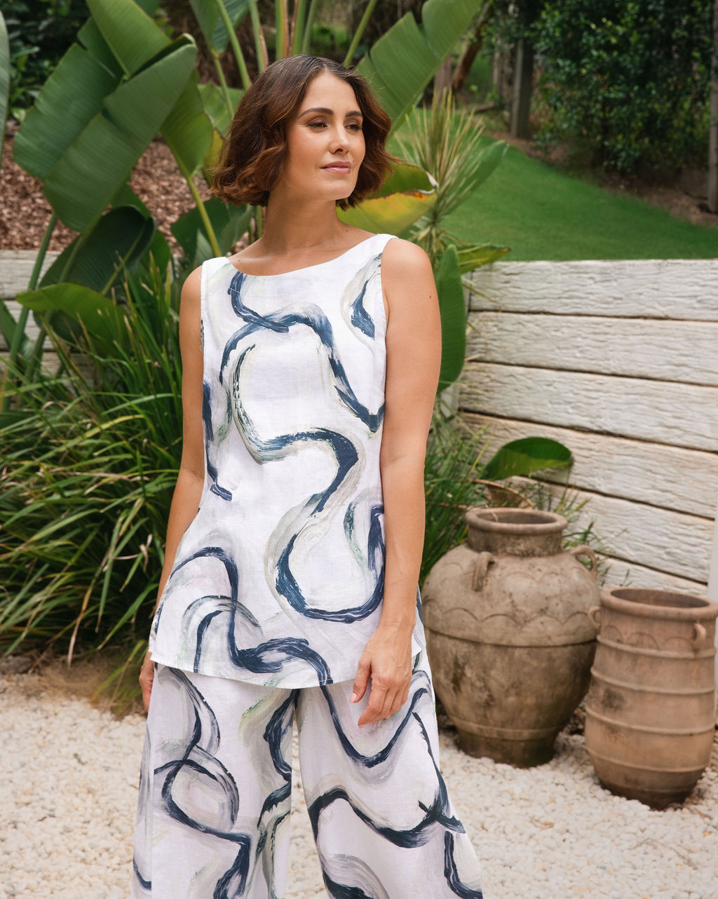A woman wearing a Riverstone sleeveless white top and matching See Saw wide-leg pants with an abstract blue and gray brushstroke pattern, standing outdoors in a garden setting with large green plants, terracotta pots, and a light stone wall.
