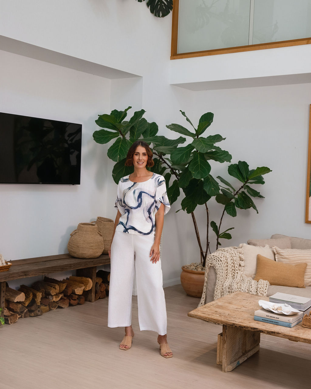 A woman wearing a white See Saw short-sleeve blouse with a blue riverstone brushstroke pattern, paired with white pants, standing in front of a potted fiddle-leaf fig plant.