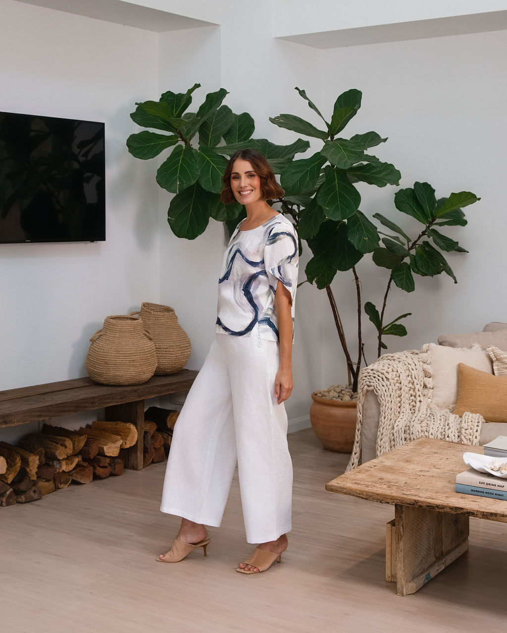 A woman wearing a white See Saw short-sleeve blouse with a blue riverstone brushstroke pattern, paired with white pants, standing in front of a potted fiddle-leaf fig plant.