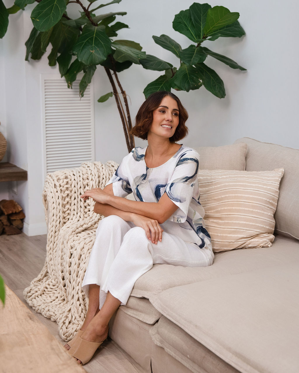 A woman wearing a white See Saw short-sleeve blouse with a blue riverstone brushstroke pattern, paired with white pants, standing in front of a potted fiddle-leaf fig plant.