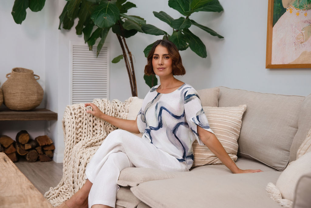 A woman wearing a white See Saw short-sleeve blouse with a blue riverstone brushstroke pattern, paired with white pants, standing in front of a potted fiddle-leaf fig plant.