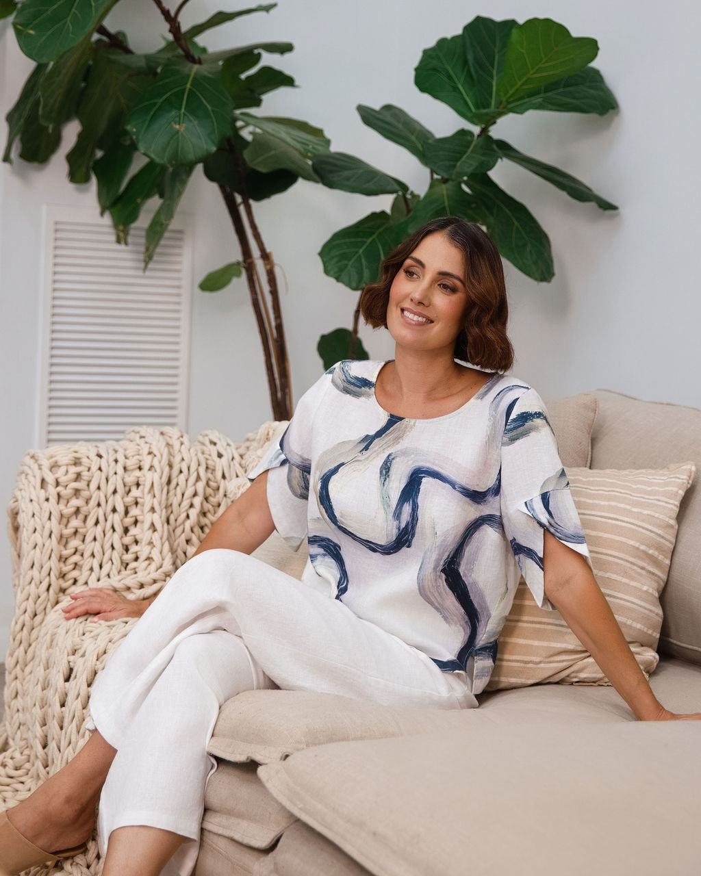 A woman wearing a white See Saw short-sleeve blouse with a blue riverstone brushstroke pattern, paired with white pants, standing in front of a potted fiddle-leaf fig plant.