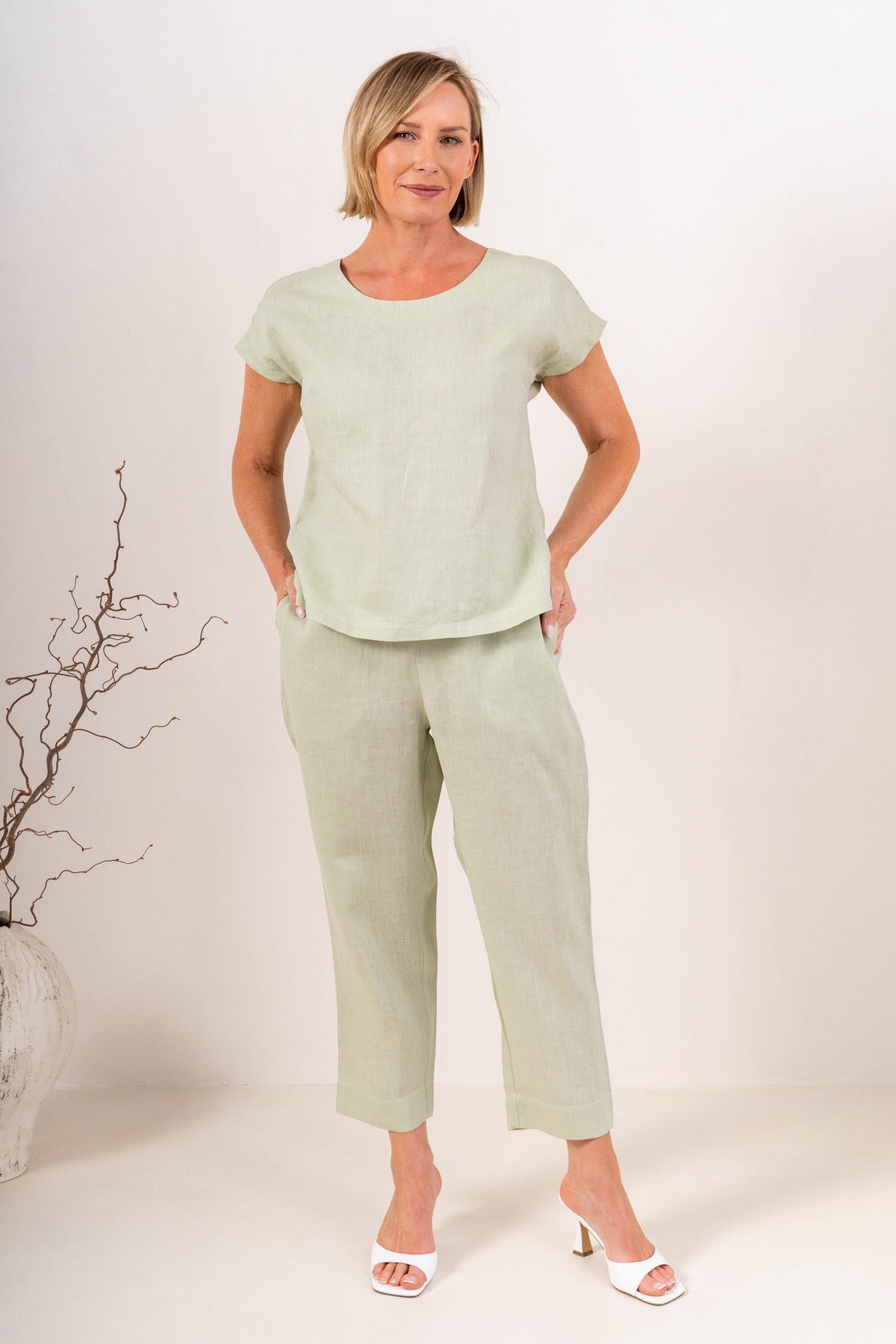 A person wearing light sage linen  See Saw 7/8 pants with a relaxed straight-leg fit, paired with white heeled slide sandals, photographed against a plain light beige studio background.
