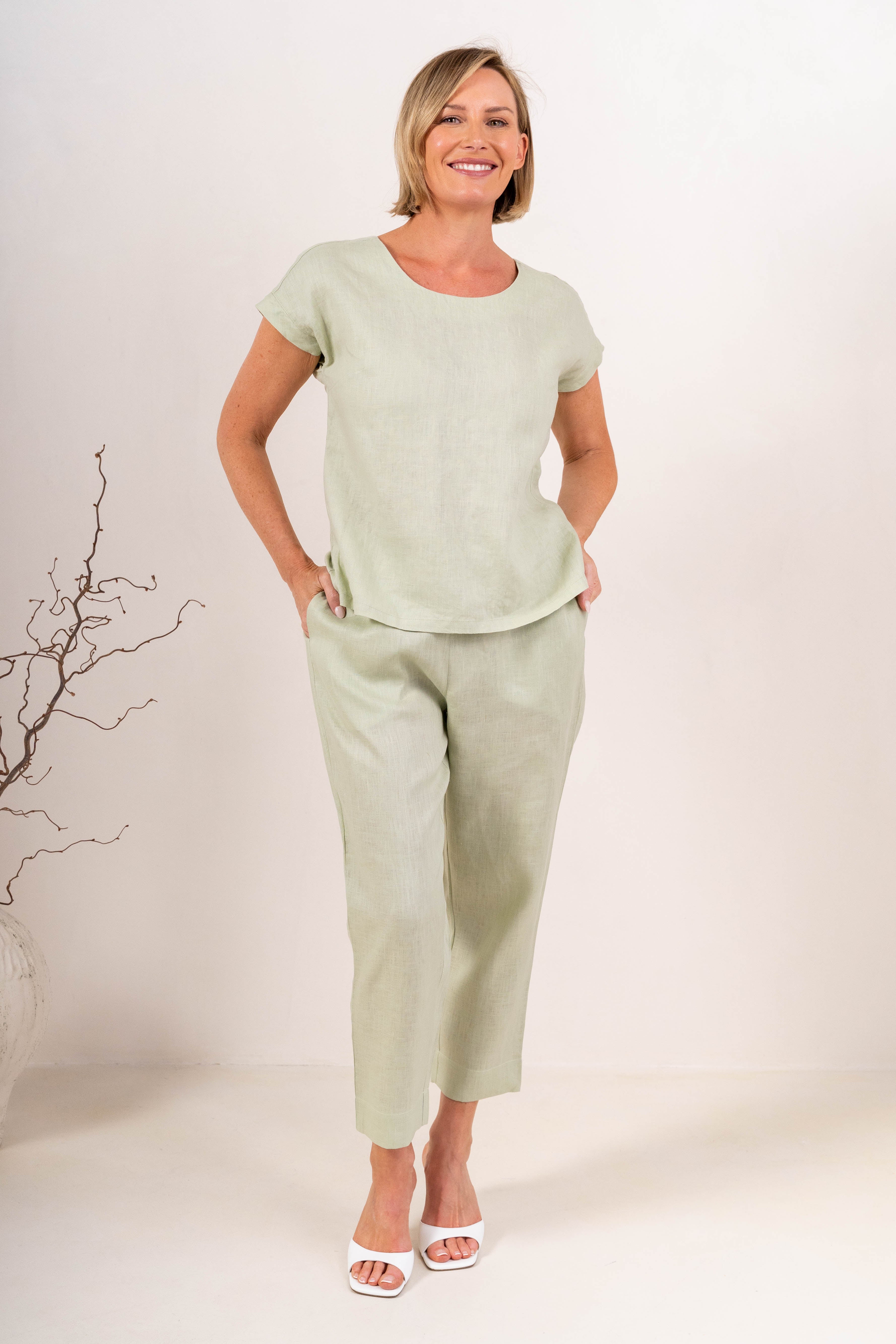 A woman wearing a light sage green See Saw linen short-sleeve top with a button-down back and matching pants, set against a plain off-white background.