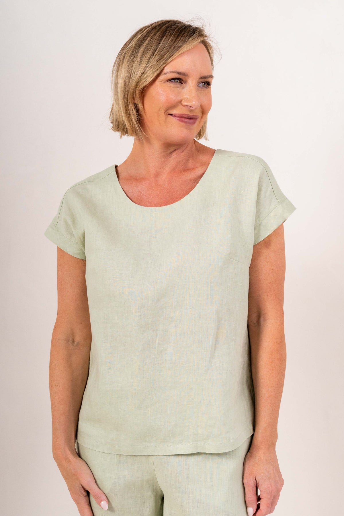 A smiling blonde woman wearing a sage short-sleeve See Saw linen top and matching marigold pants, photographed against a plain light beige studio background.