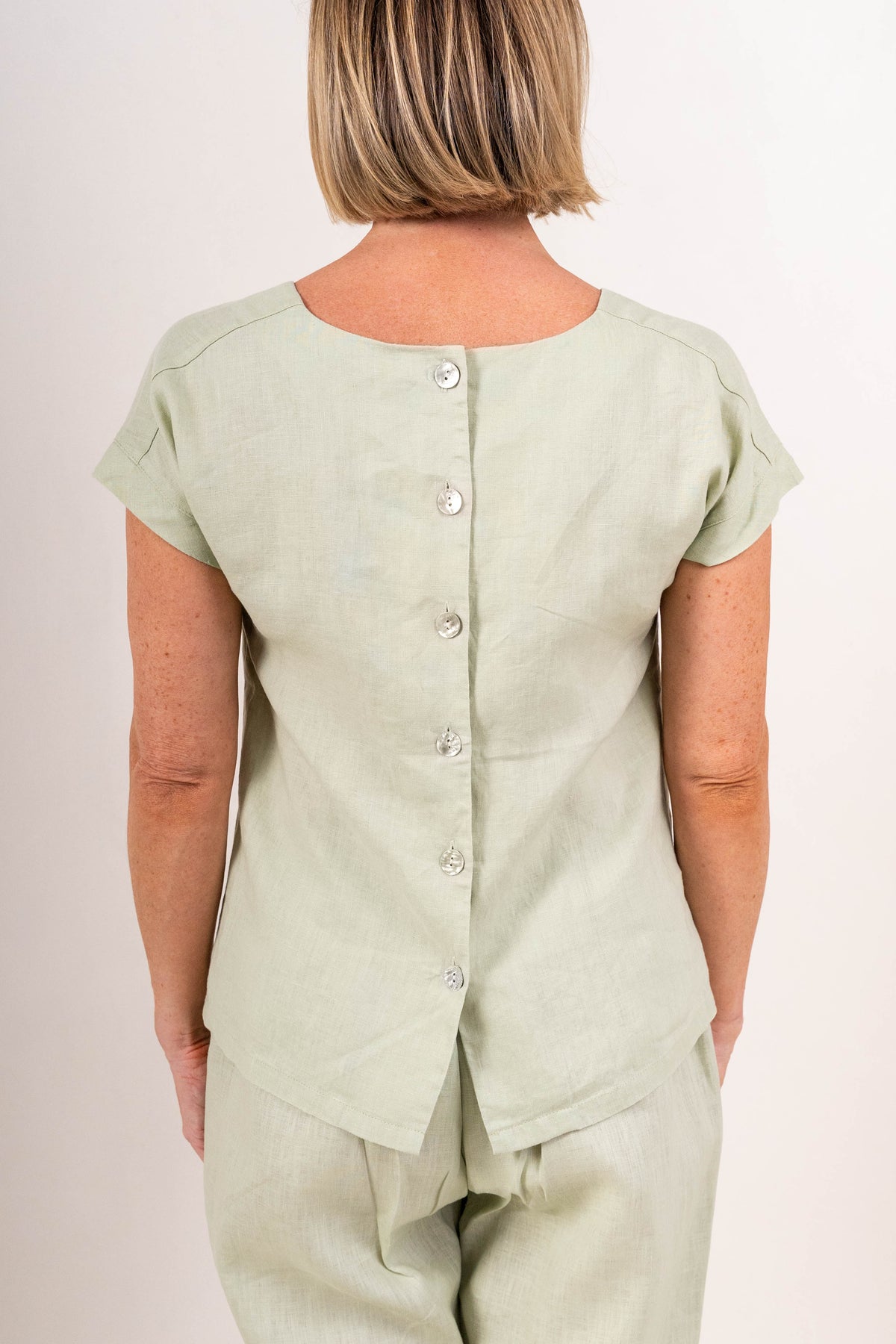 A woman viewed from the back wearing a light sage green See Saw linen short-sleeve top with a button-down back and matching pants, set against a plain off-white background.