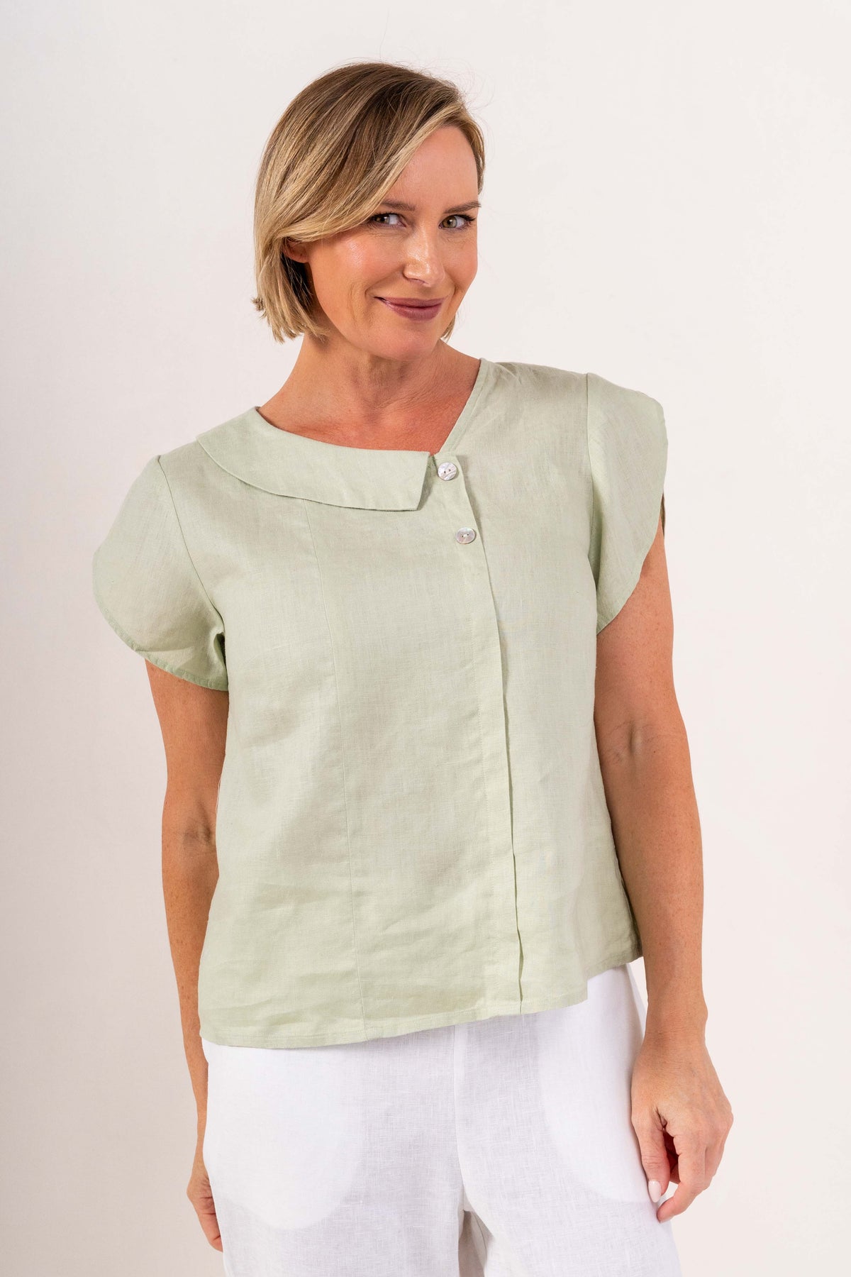 A woman modeling a sage short-sleeve button-up top and high-waisted cropped trousers with a colorful abstract floral print, standing against a plain off-white studio background.
