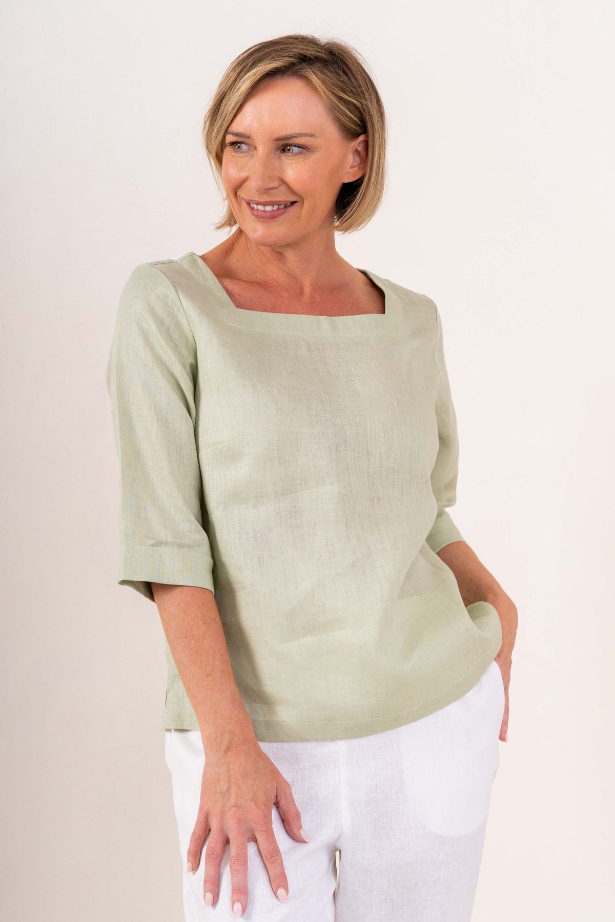 A woman wearing a light sage linen See Saw top with a square neckline and elbow-length sleeves, paired with colorful petal drift pants, standing in a studio.