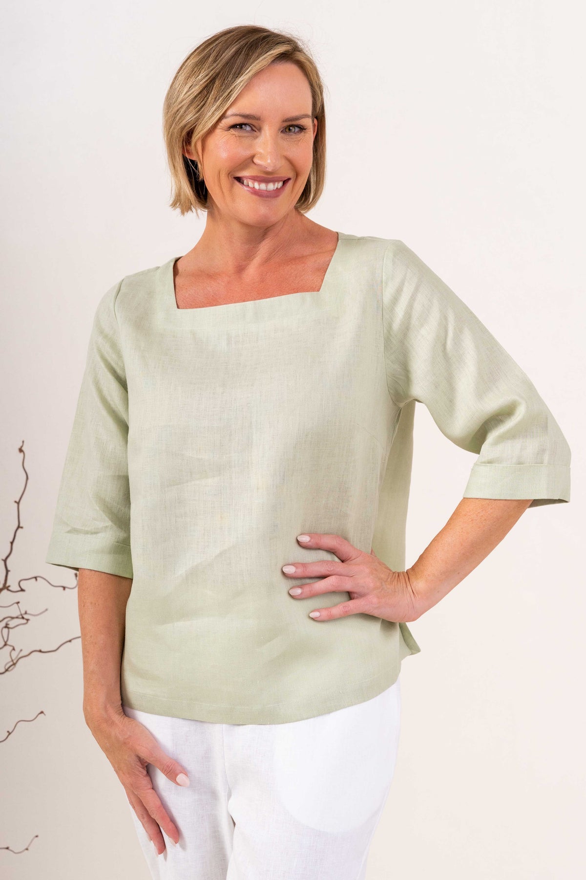 A woman wearing a light sage linen See Saw top with a square neckline and elbow-length sleeves, paired with colorful petal drift pants, standing in a studio.