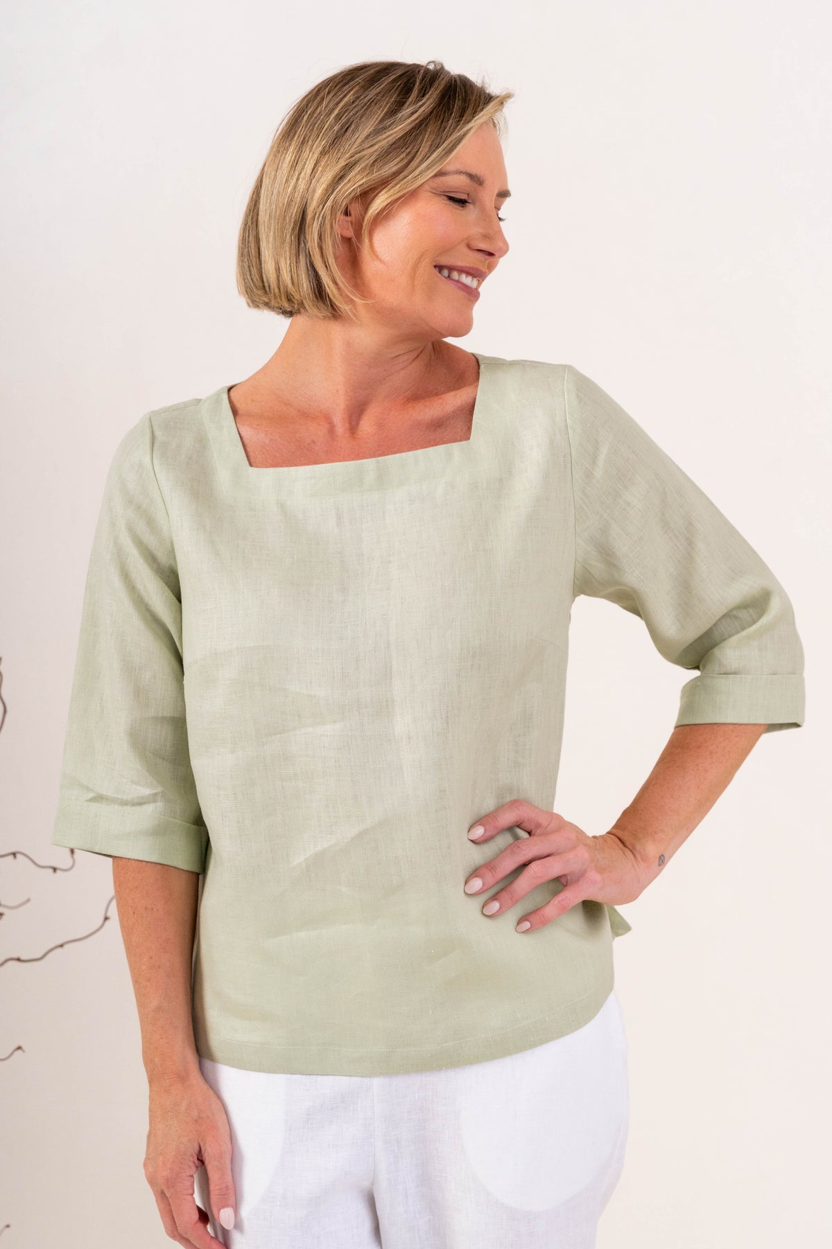 A woman wearing a light sage linen See Saw top with a square neckline and elbow-length sleeves, paired with colorful petal drift pants, standing in a studio.