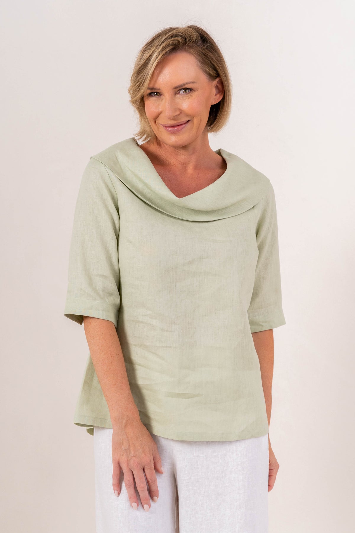 A woman modeling a sage linen See Saw top with a wide draped collar and elbow-length sleeves, paired with white pants, against a plain off-white background.