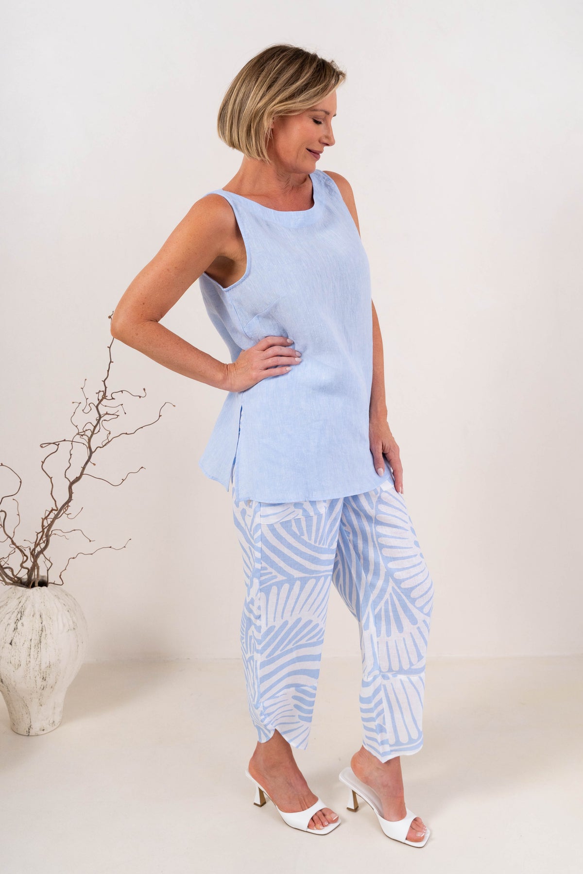 A woman modeling a light blue and white Palm Breze See Saw Linen two-piece set consisting of a short-sleeve top and matching pants, standing against a plain off-white studio background.