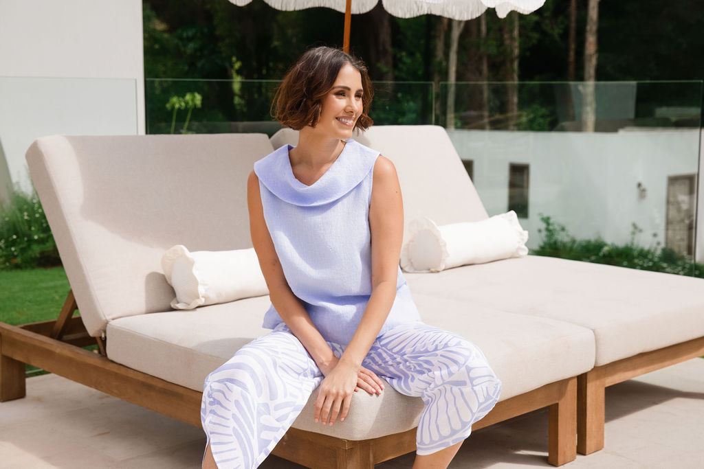 A woman modeling a sky blue sleeveless cowl-neck top and matching light blue and white palm breeze See Saw print wide-leg pants, standing in a modern outdoor patio setting with a lounge chair and white umbrella.