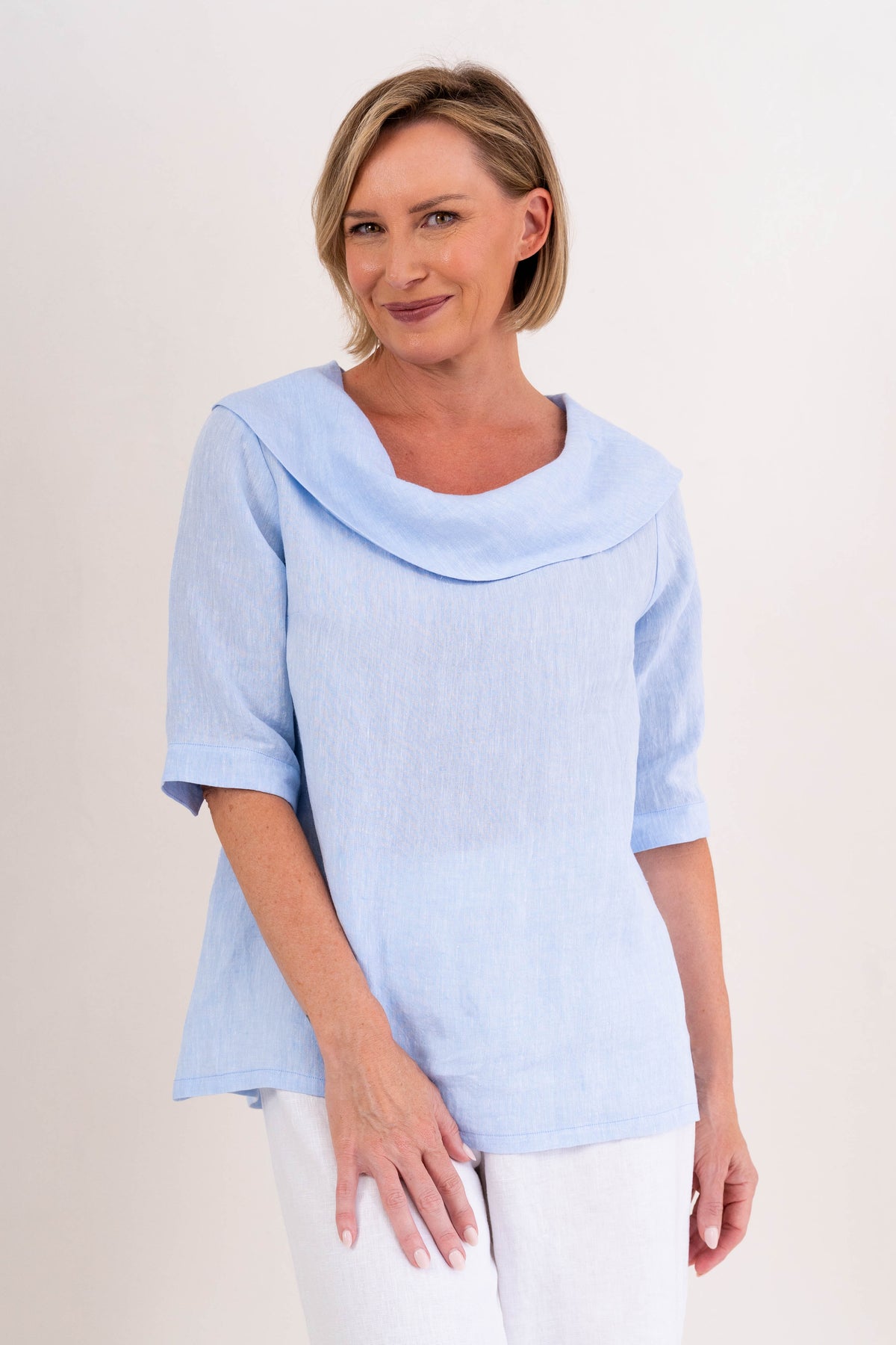 A woman modeling a sky blue linen See Saw top with a wide draped collar and elbow-length sleeves, paired with white pants, against a plain off-white background.