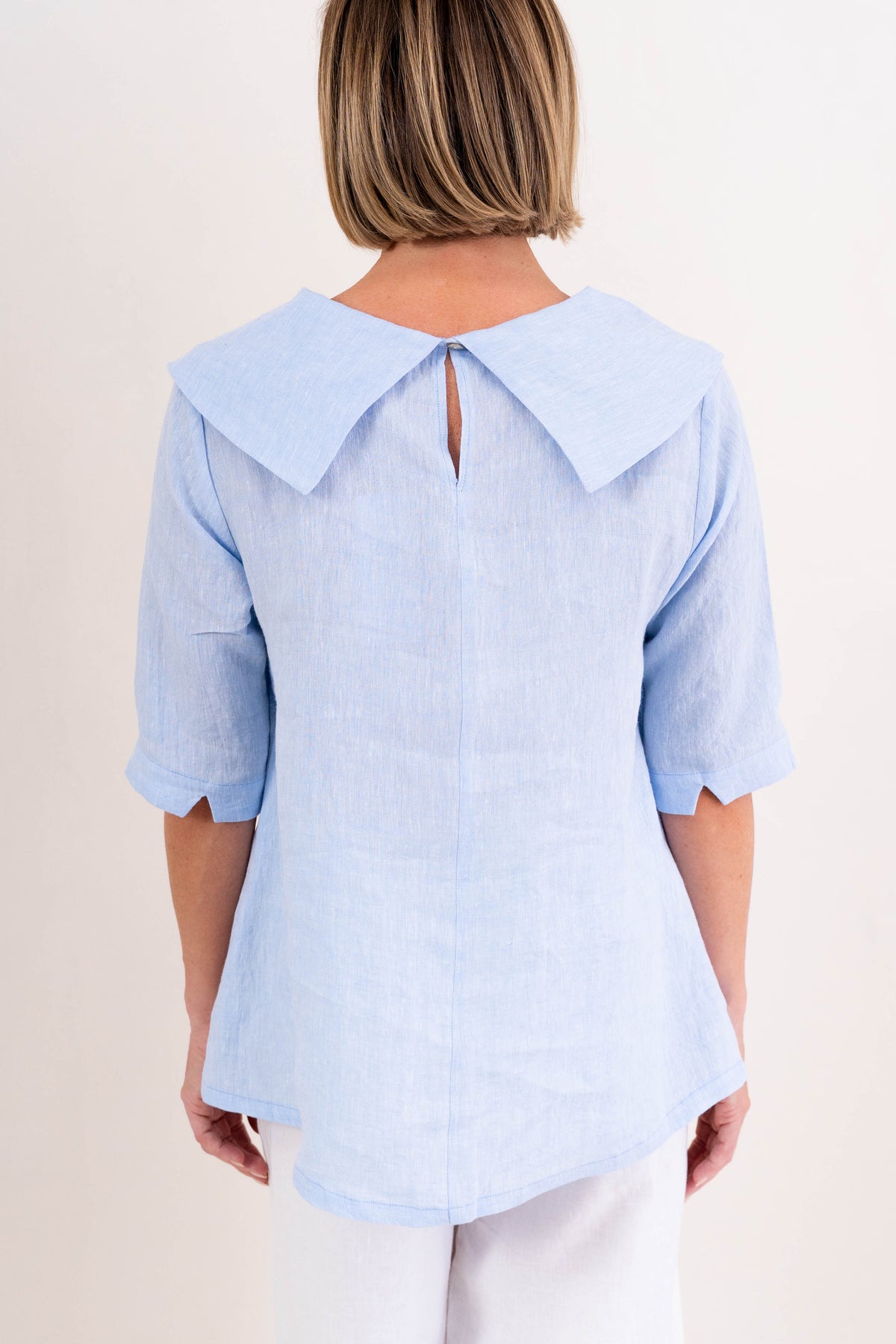 A woman modeling a sky blue linen See Saw top with a wide draped collar and elbow-length sleeves, paired with white pants, against a plain off-white background.