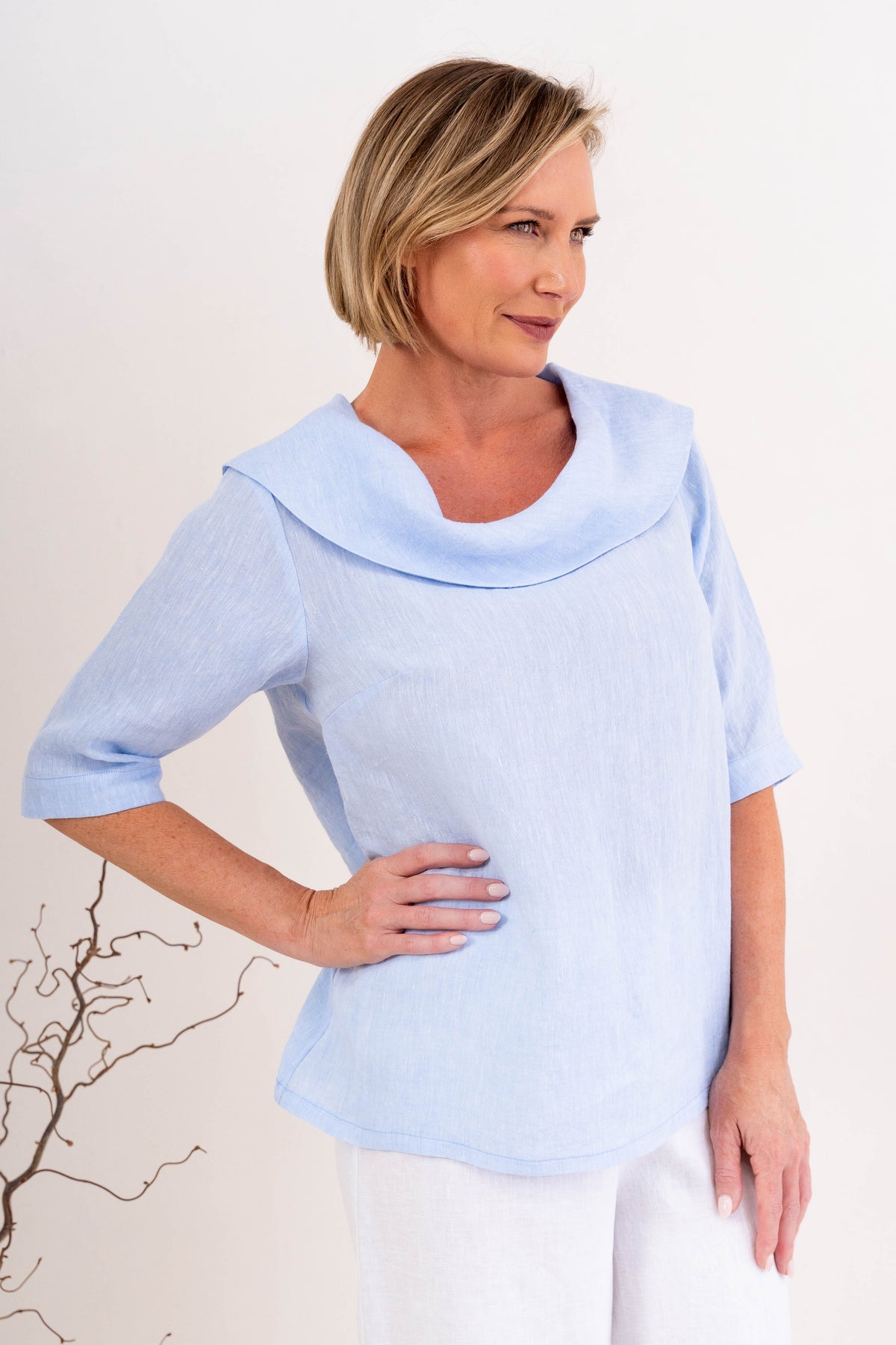A woman modeling a sky blue linen See Saw top with a wide draped collar and elbow-length sleeves, paired with white pants, against a plain off-white background.
