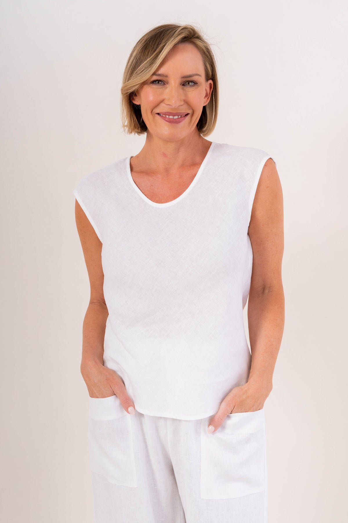 A smiling blonde woman wearing a white sleeveless V-neck top in linen by See Saw and matching white pants, standing against a plain light background.