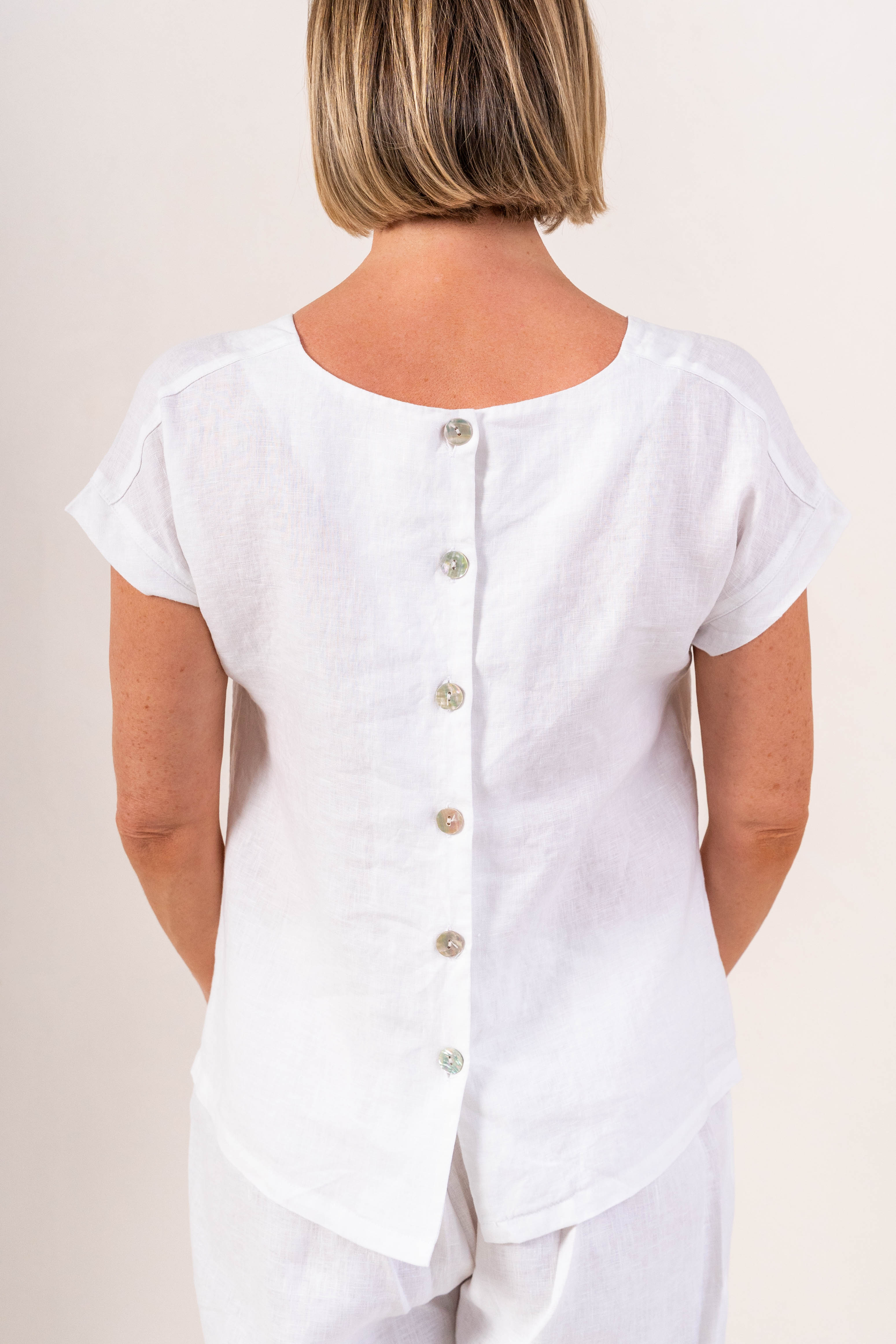 A woman wearing a white short-sleeve See Saw linen top and matching white pants, photographed against a plain light beige studio background.