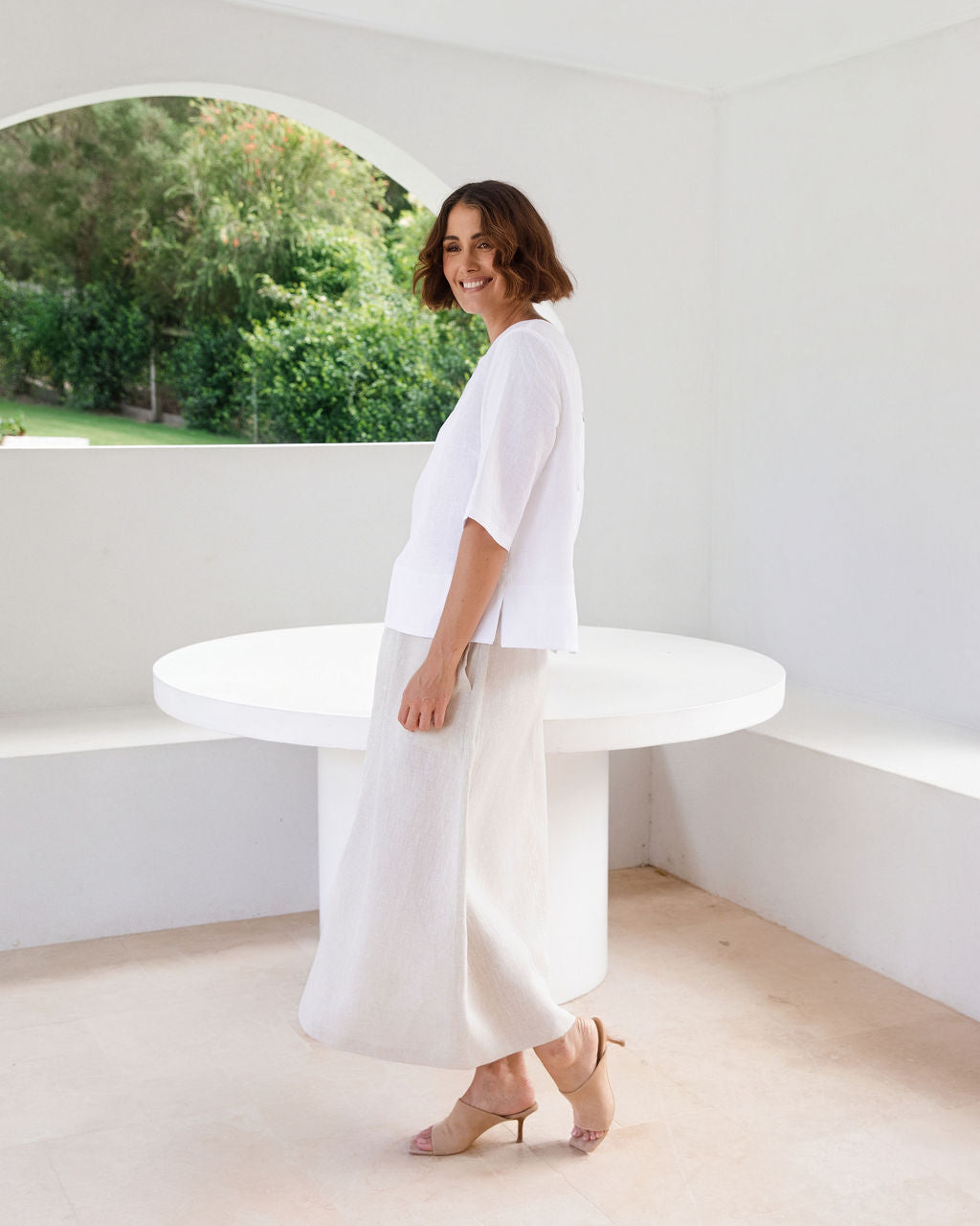 A woman wearing a See Saw white short-sleeve top and white maxi skirt stands in a bright, minimalist white room with a round white table, looking back over her shoulder. She is wearing beige high-heeled sandals.