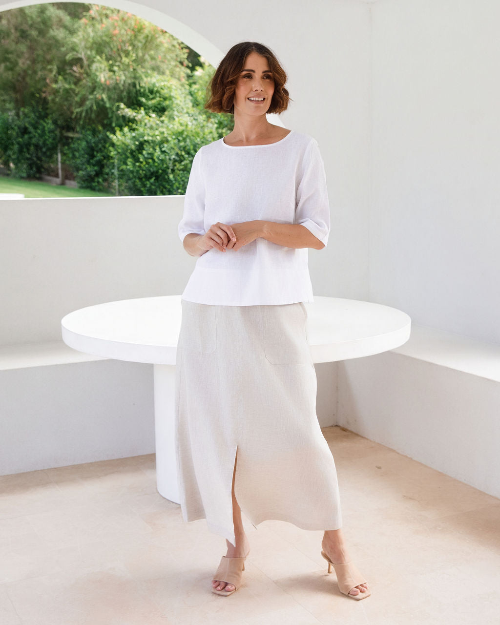 A woman modeling a white linen See Saw three-quarter sleeve top and light beige cropped trousers, standing in a bright, modern white room with a round white table.