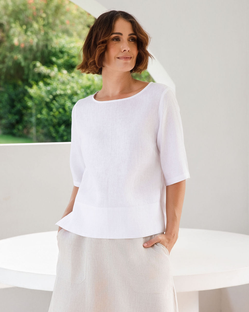 A woman wearing a white linen See Saw short-sleeve linen top and a light beige linen skirt, standing in a bright, modern white architectural setting with green foliage visible in the background.
