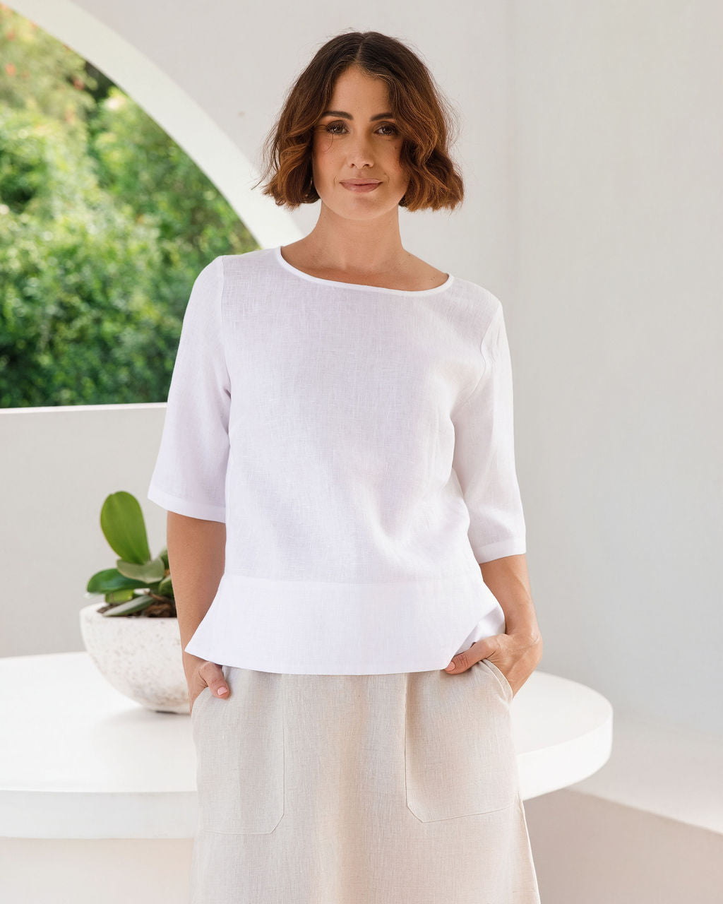A woman wearing a white linen See Saw short-sleeve knit top with a peplum hem and light beige linen trousers, standing in a bright, modern white room with an arched window view of green foliage.