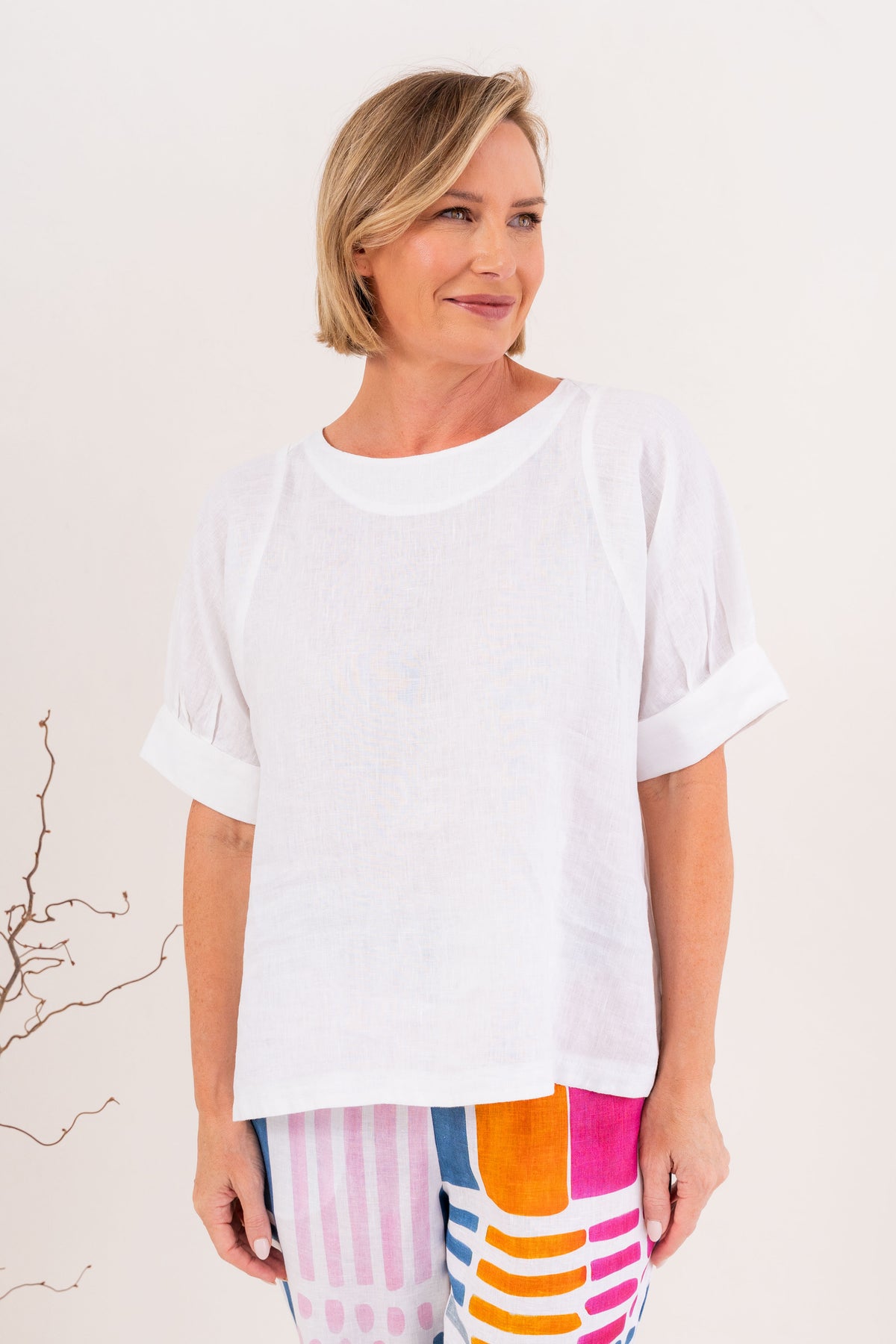 A woman wearing a white linen See Saw short-sleeve top with rolled cuffs and a relaxed fit, paired with colorful striped pants, standing against a plain light background.