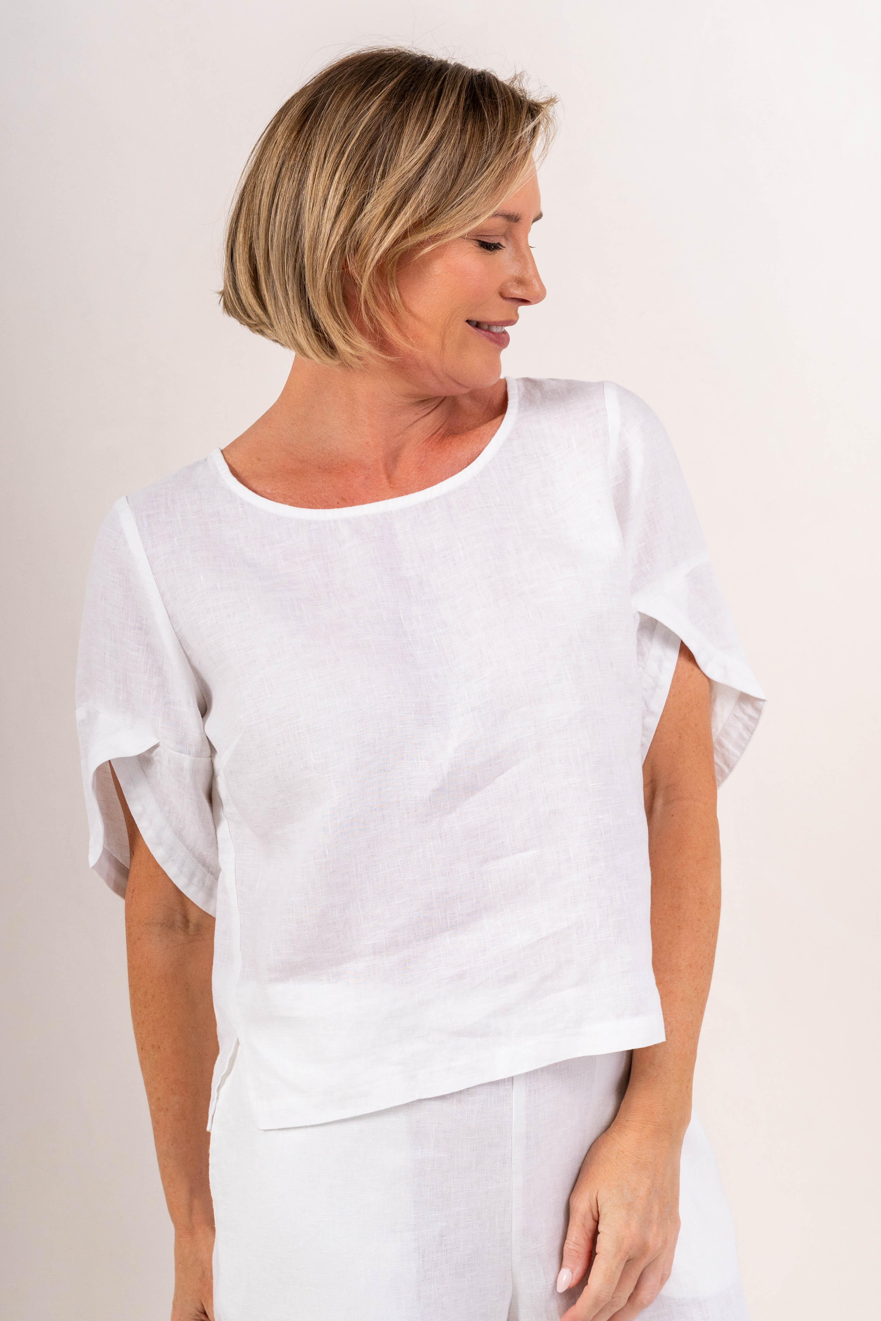 A woman wearing a white See Saw linen short-sleeve top with ruffled or layered sleeve details and white pants, photographed against a plain light background.
