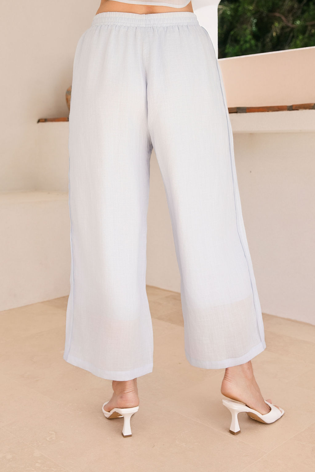 A woman modeling a See Saw Ramie blue mist  wide-leg cropped pants, standing in a bright white architectural interior with an arched window showing greenery outside