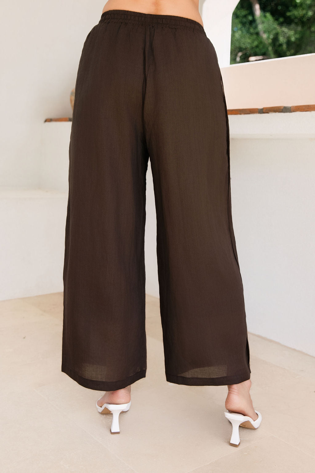 A person shown from the back wearing Ramie See Saw loose, wide-leg cropped pants in a espresso color, paired with white heeled sandals in a bright indoor setting.