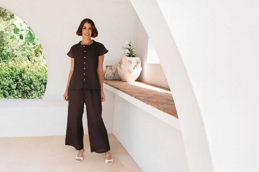 A woman modeling a dark espresso See Saw Ramie two-piece outfit consisting of a short-sleeve button-front top with draped sleeves and matching wide-leg pants, standing in a bright white architectural setting.
