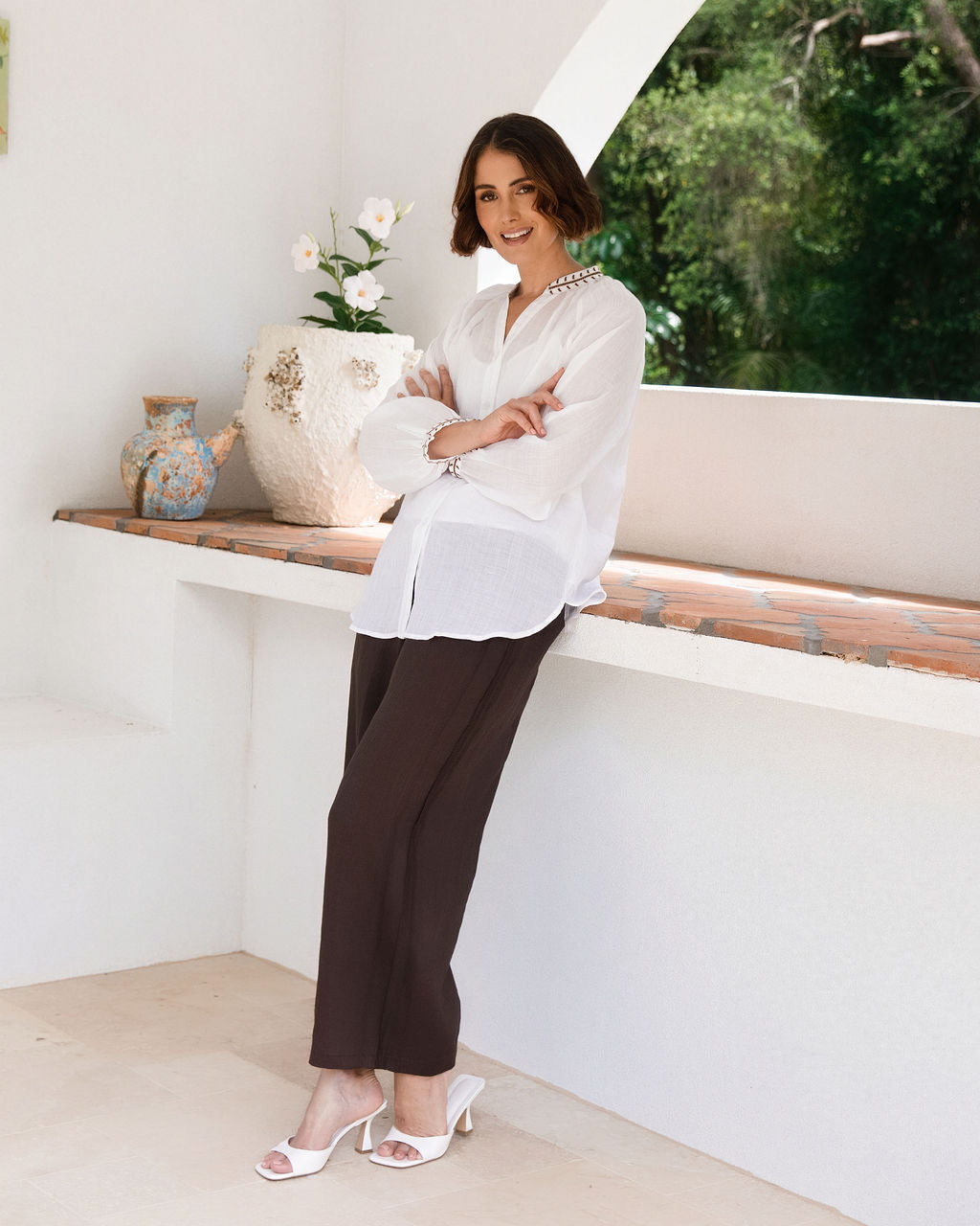 A woman wearing a white Ramie See Saw long-sleeve blouse and dark brown wide-leg pants leans against a white architectural wall with a tiled bench, vases, and greenery in the background.