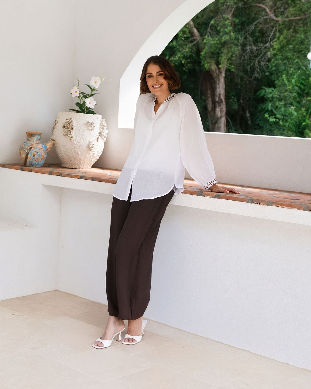 A woman wearing a white See Saw Ramie long-sleeve button-up blouse and dark brown wide-leg trousers, leaning against a white wall in a sunlit architectural setting with a window view of green trees.