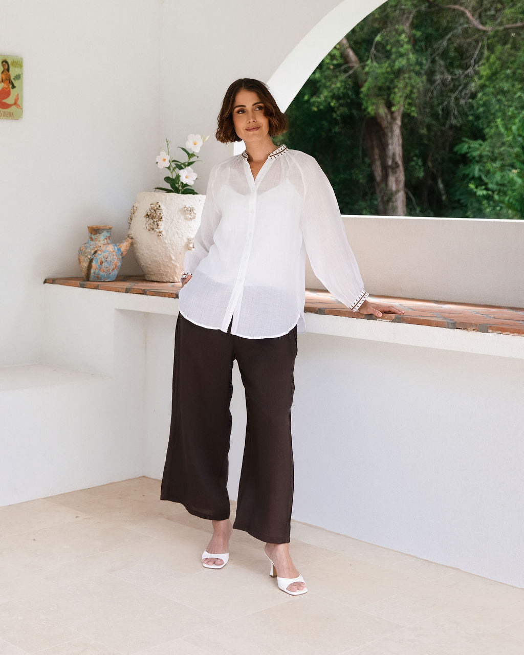 A woman modeling a white See Saw Ramie button-front top and espresso wide-leg pants, standing in a bright white architectural interior with a tiled ledge and potted plant.