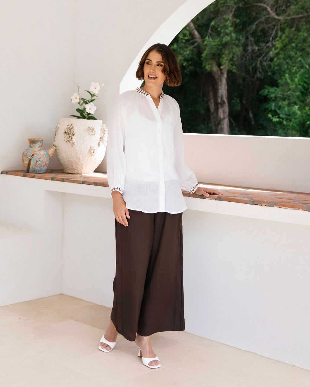 A woman modeling a white Ramie See Saw long-sleeve button-up blouse with ruffled cuffs and dark brown wide-leg pants, standing in a bright, modern white architectural setting with a view of green trees in the background.