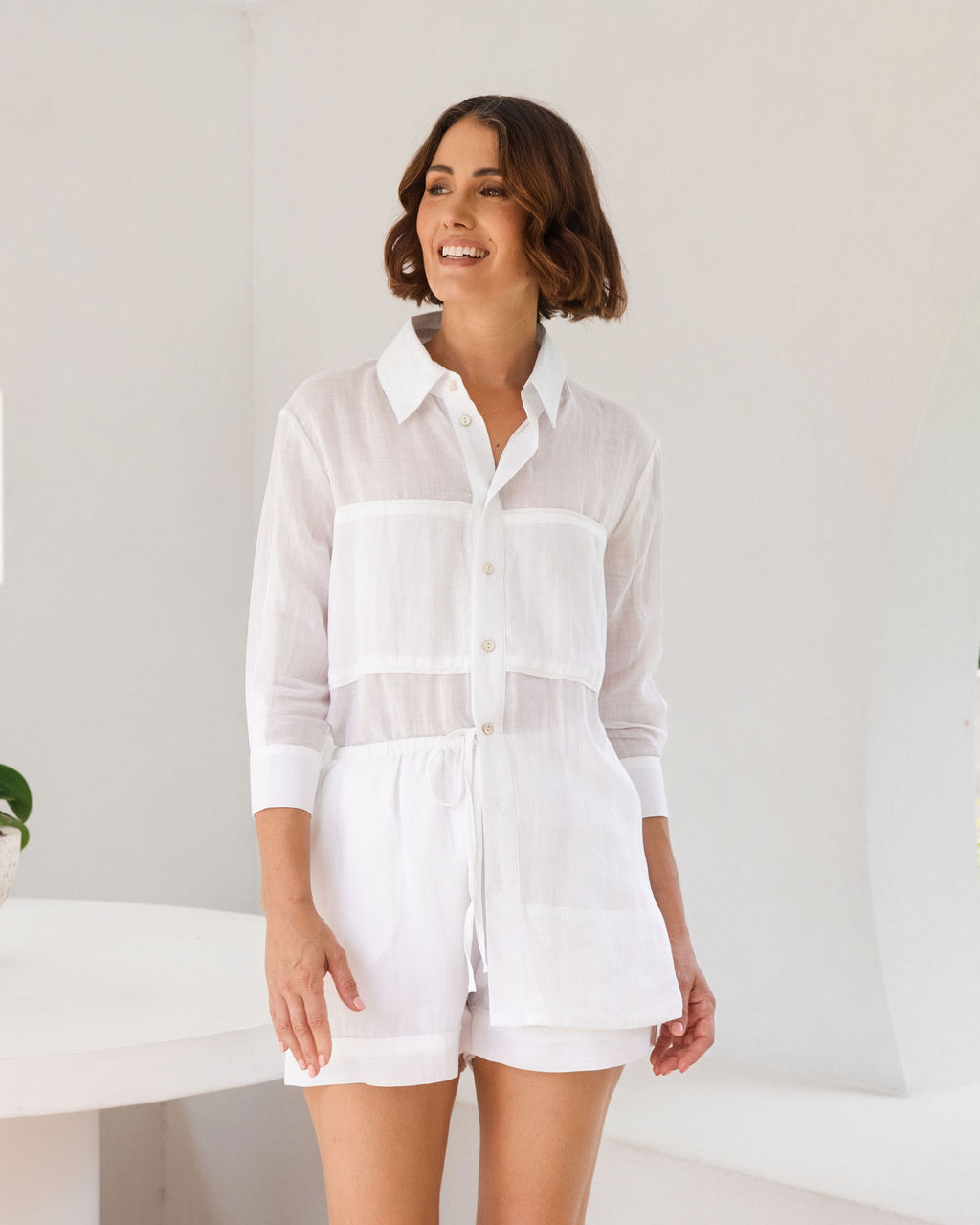 A woman wearing a fresh white see saw ramie long-sleeve shirt sits on a white architectural ledge with terracotta tile trim, with lush green foliage visible in the background.