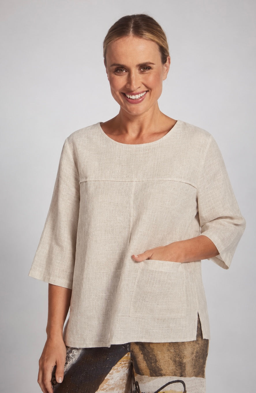 A woman modeling a natural linen See Saw top with three-quarter sleeves and a front pocket, set against a neutral gray studio background.