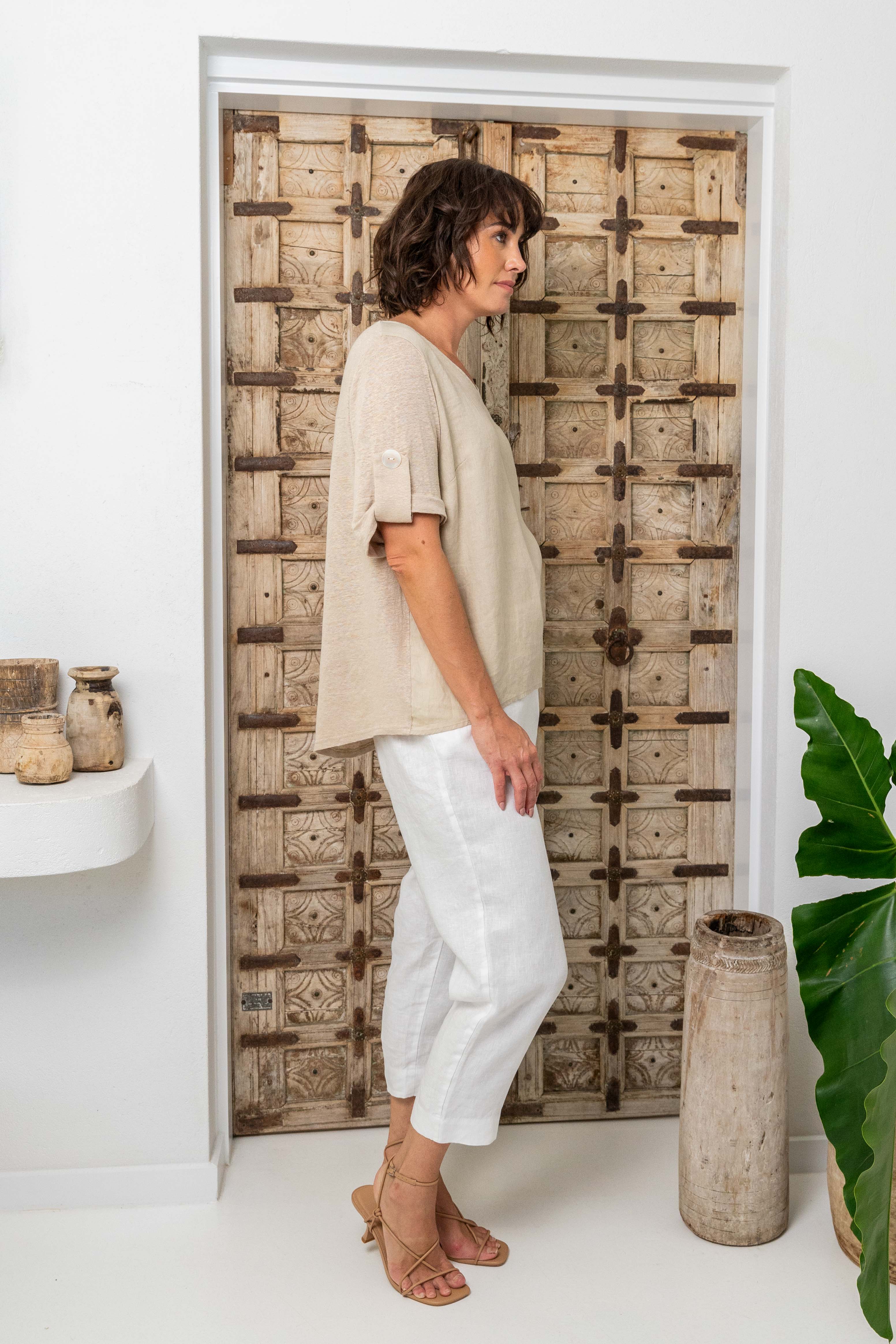 Woman wearing a beige top and white pants standing against a wooden wall. See Saw Mixed Linen Blend T-shirt