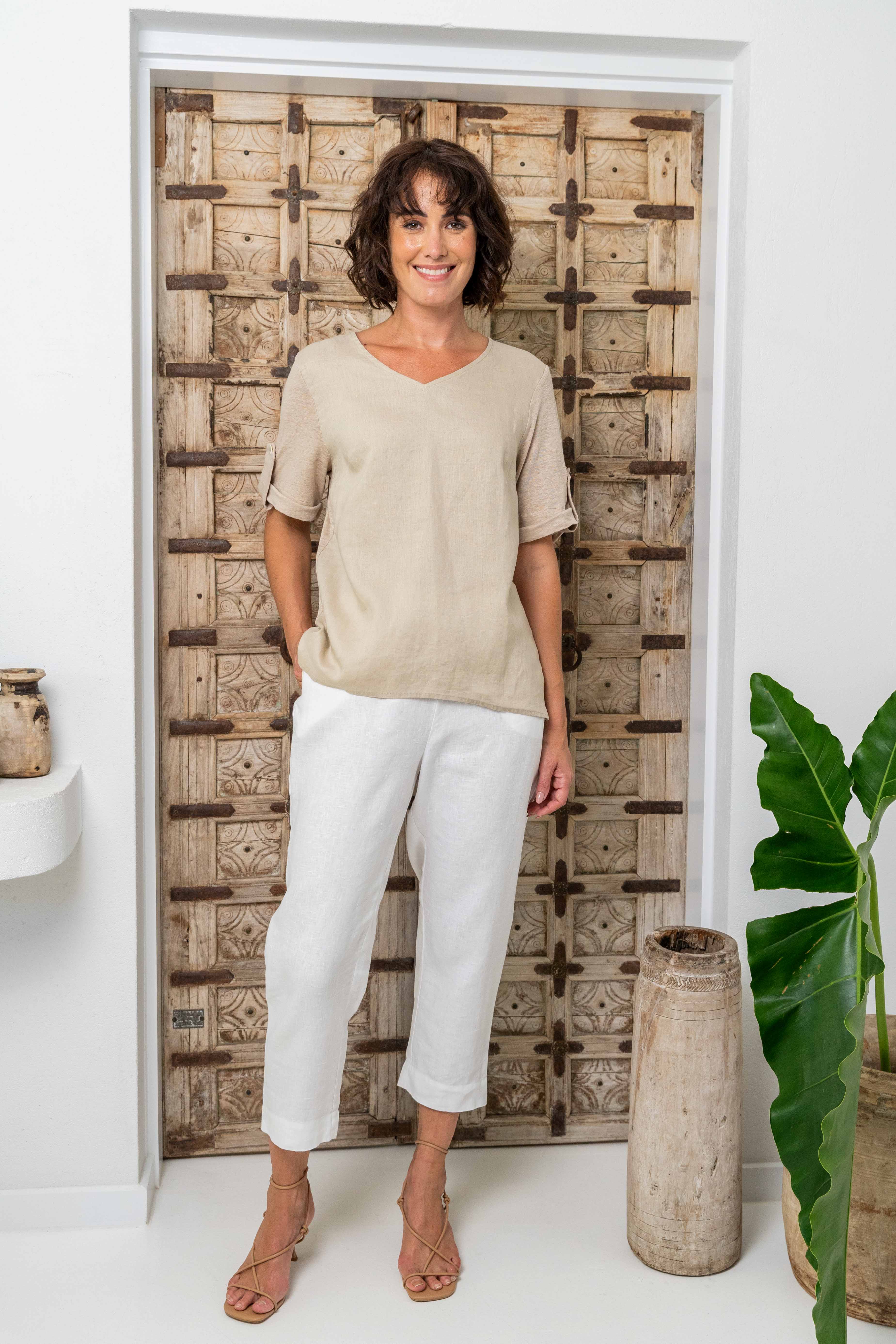 Woman wearing a beige top and white pants standing against a wooden wall. See Saw Mixed Linen Blend T-shirt