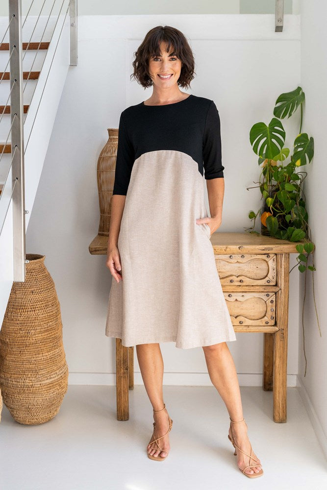 Woman wearing a rayon and linen See Saw dress standing in front of a white background and drawers and a plant. 