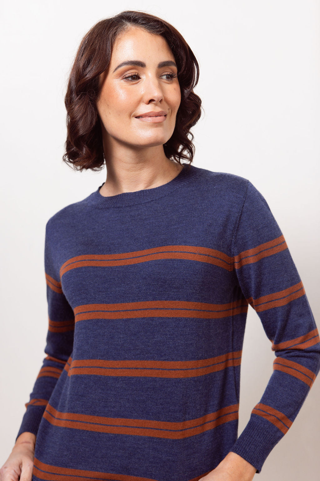 Woman wearing a indigo See Saw Merino wool sweater with copper stripes and dark jeans against a plain background