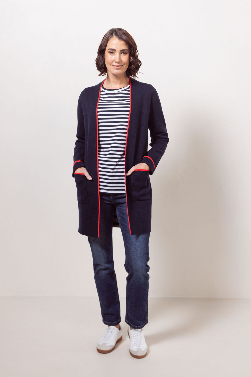 Woman wearing a See Saw navy cardigan with red trim, striped shirt, jeans, and white sneakers on a plain background