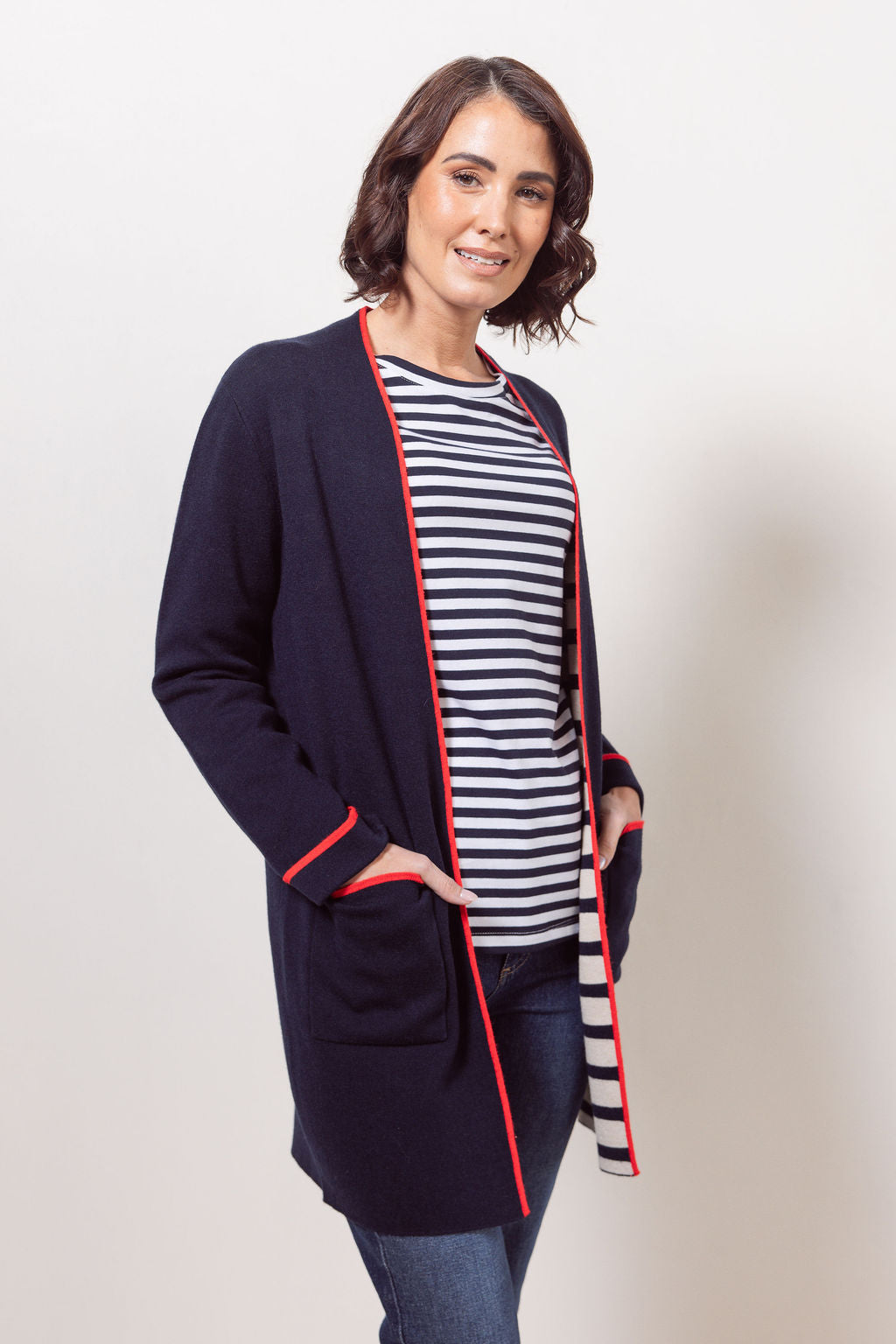 Woman wearing a See Saw navy cardigan with red trim, striped shirt, jeans, and white sneakers on a plain background