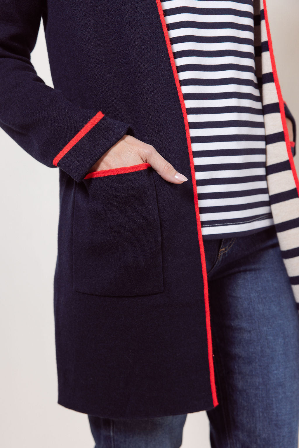 Woman wearing a See Saw navy cardigan with red trim, striped shirt, jeans, and white sneakers on a plain background