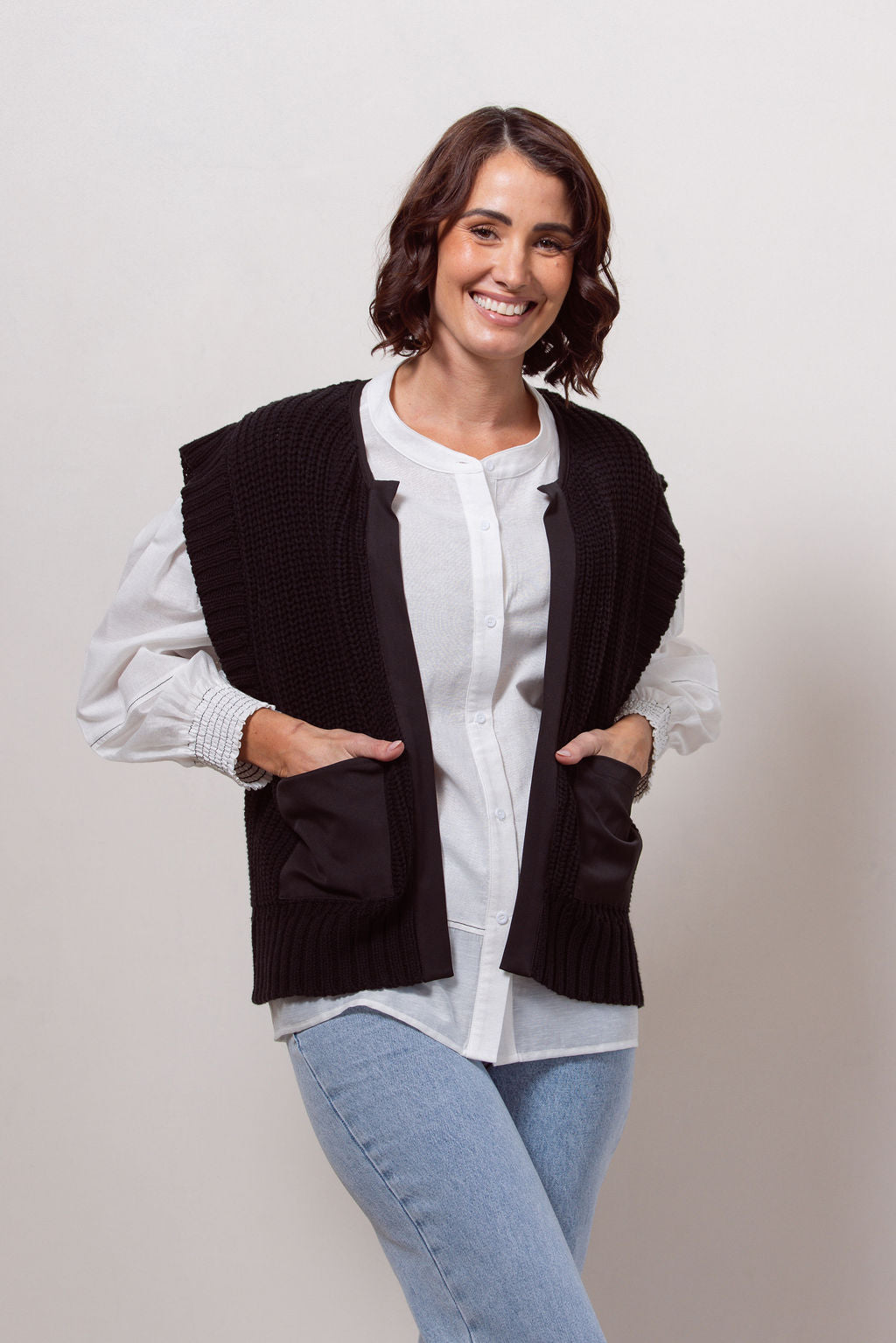 Woman wearing a black cotton See Saw knitted vest over a white shirt and blue jeans against a white wall.