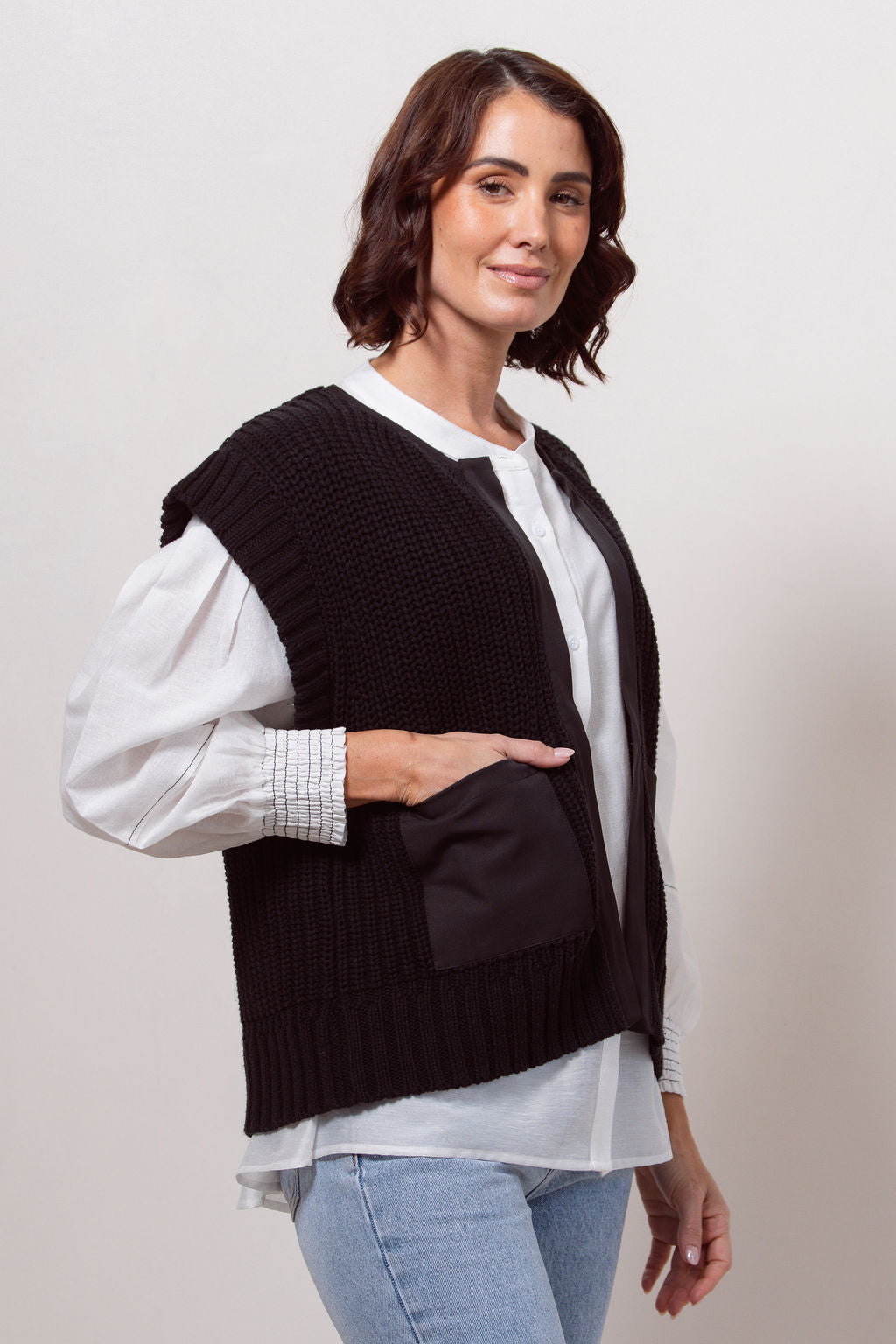 Woman wearing a black cotton See Saw knitted vest over a white shirt and blue jeans against a white wall.