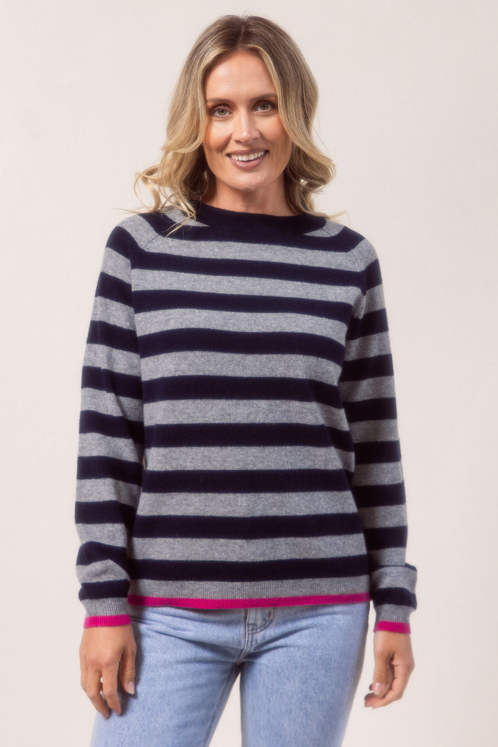 Woman wearing a Merino Wool See Saw navy and grey striped sweater with light blue jeans on a white background